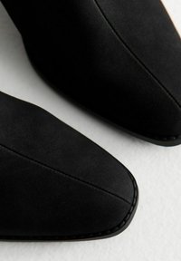 Botas de tobillo de ante negro con punta afilada, que presentan costuras bordadas y una textura suave, sobre un fondo claro.
