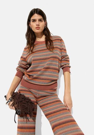 Knitted striped sweater in rust, gray, and cream; relaxed fit with ribbed cuffs. Model holds a brown fringed clutch bag.