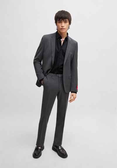 Сив tailored костюм с черна риза с копчета. Забележителен червен акцент на ръкава на якето. Моделът носи черни обувки с метални детайли.
