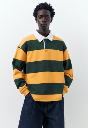 Młody mężczyzna z krótkimi kręconymi włosami nosi żółtą i ciemnozieloną koszulkę rugby w szerokie paski z białym kołnierzem oraz ciemnoniebieskie dżinsy.