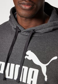Sudadera con capucha gris, hecha de una tela suave, que presenta un logo de Puma en blanco saltando y cordones en contraste de color negro en la capucha.