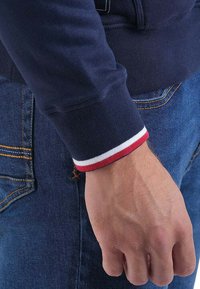 Sweat-shirt bleu marine avec des poignets ajustés ornés de rayures rouges et blanches. Porté avec un jean bleu, montrant un gros plan du poignet.