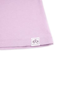 Ljust lila bomull t-shirt nederkant med en liten vit etikett som har lila grafisk design och initialerna BXR. Slät textur, avslappnad stil.