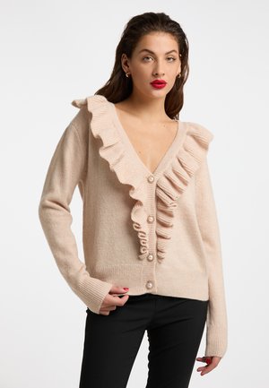 Frau mit langen braunen Haaren, die eine hellrosa, geraffte Strickjacke mit Perlenknöpfen und schwarze Hosen vor weißem Hintergrund trägt.