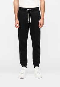 Μαύρο παντελόνι φόρμας με ελαστική μέση, κορδόνι ρύθμισης, πλαϊνές τσέπες και στενή εφαρμογή, συνδυασμένο με λευκά αθλητικά παπούτσια.