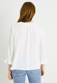 Blusa blanca con un cuello con volantes, mangas tres cuartos y un corte relajado, combinada con jeans azules. La tela parece suave y ligera.