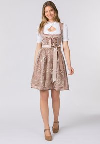 Rosa gemusterter Dirndl mit Spitzendetails, taillierter Bluse und Schleife in der Taille. Kombiniert mit einer weißen Bluse und braunen Schuhen.