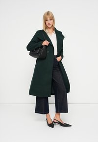 Sötétzöld gyapjú kabát széles gallérral, gombos záródással, fehér inggel, magas derekú sötét farmer széles szárú nadrággal és fekete magas sarkú slingback cipővel.