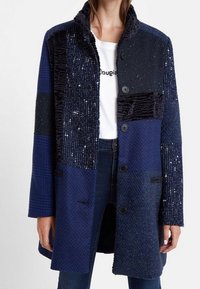 Marinblå lapptäcktsjacka med olika texturer, inklusive velour och vävda mönster, med fem knappar och en designs som krage.