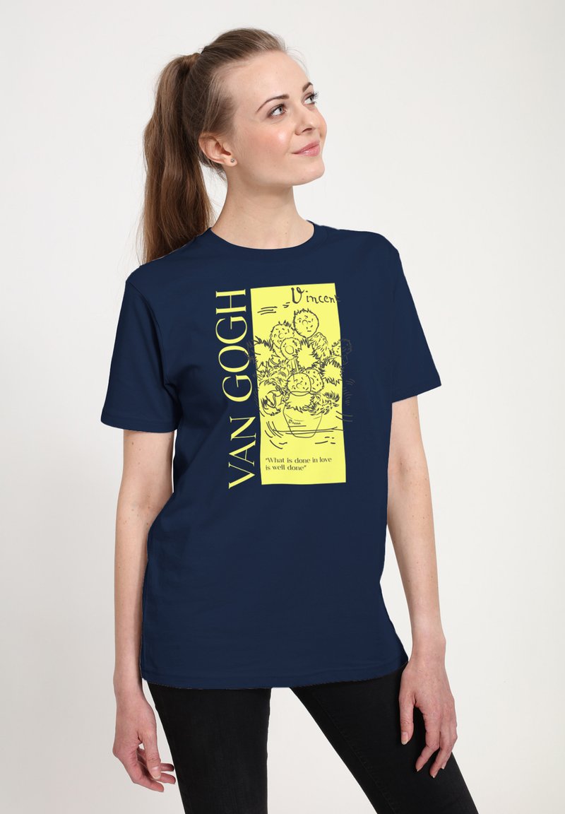 Donna con maglietta blu navy con schizzo giallo di girasoli di Van Gogh e citazione, in piedi con la testa inclinata e un leggero sorriso.