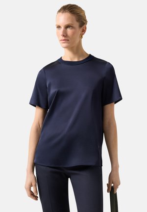 Kobieta o jasnej karnacji i blond włosach ubrana w granatowy satynowy top z krótkim rękawem i spodnie, trzymająca czarny pasek w jednej ręce, patrząca lekko w prawo.