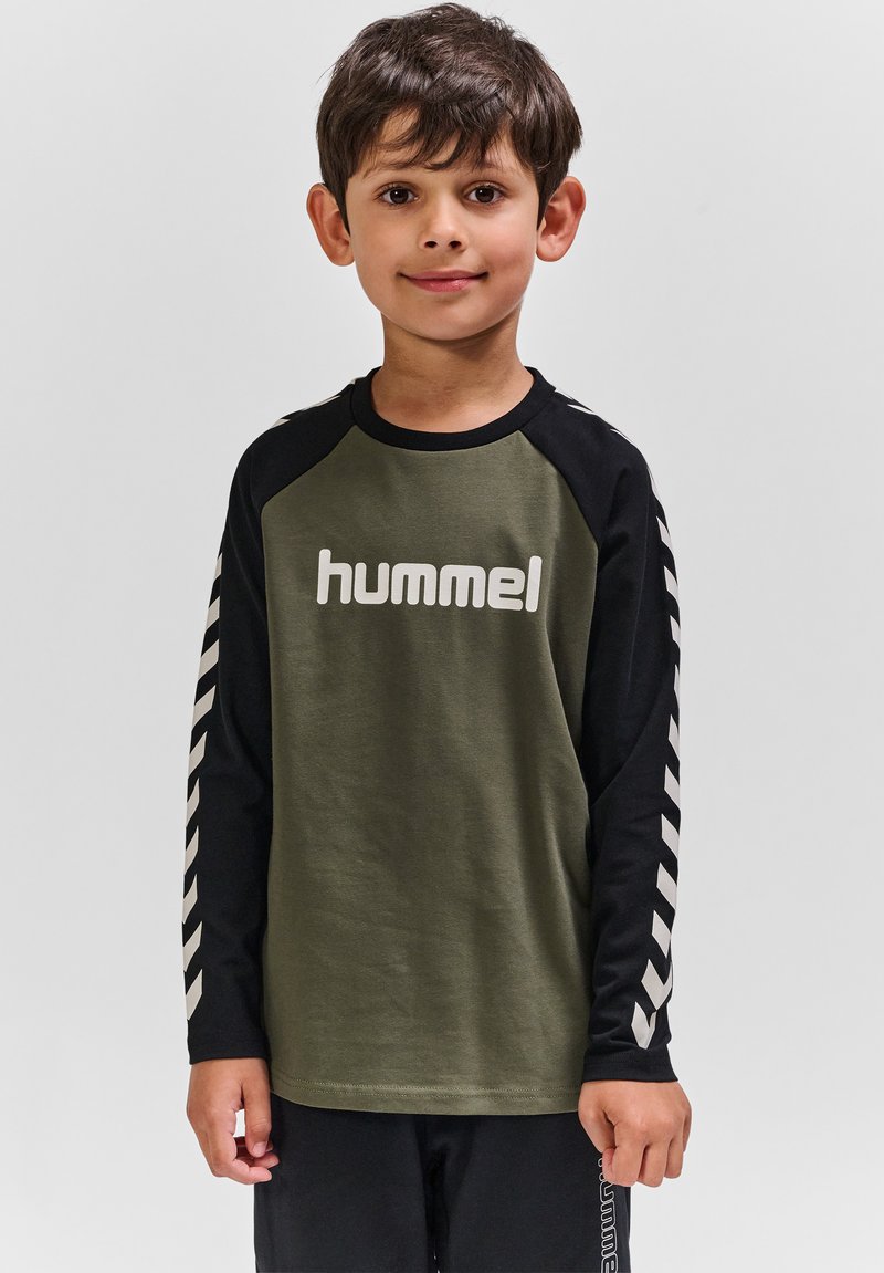 Junger Junge steht und schaut nach vorne, trägt ein olivfarbenes und schwarzes Langarmshirt mit dem "hummel"-Logo und Chevron-Mustern an den Ärmeln.