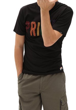 Personne portant un t-shirt noir avec le texte coloré « PRIDE » et un short cargo vert olive, une main dans la poche, l'autre près du visage.