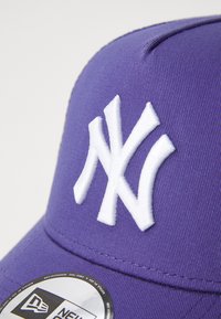 Lilla baseballcap lavet af tekstureret stof, med et fremtrædende hvidt broderet New York Yankees-logo på forsiden og et mærket rundt klistermærke.