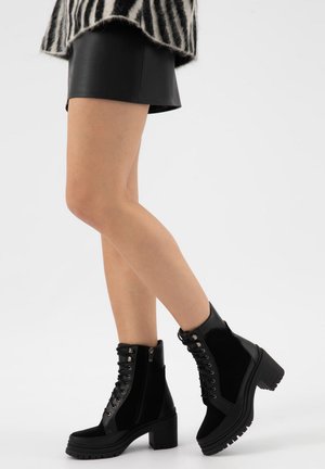 Beine mit schwarzen, klobigen, geschnürten Stiefeletten mit Reißverschluss, kombiniert mit schwarzen Leder-Shorts und einem schwarz-weiß gestreiften Oberteil.