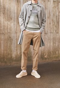 Cappotto lungo grigio, maglione verde a trama, chinos beige e sneakers crema. Look a strati con colori contrastanti e toni neutri. Superfici lisce e vestibilità casual.