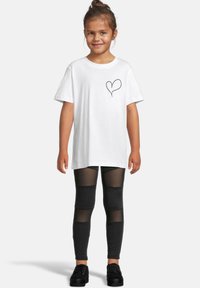 Hvit bomull T-skjorte med svart hjertegrafikk på brystet, kombinert med svarte tights med gjennomsiktige paneler. Svarte sko fullfører antrekket.