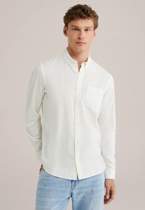 Chemise blanche à manches longues et à boutons avec un motif texturé et une poche poitrine, assortie à un jean bleu clair.