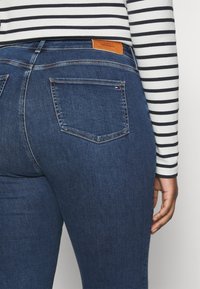 Tmavě modré denimové džíny s dvěma zadními kapsami a hnědou koženou nášivkou. Pruhované tričko s dlouhým rukávem viditelné nad páskem.