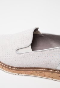 Chaussure blanche à enfiler avec des points irisés, un panneau latéral élastique, un intérieur en cuir rembourré et une semelle en liège texturée avec une bande de roulement blanche.