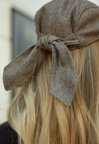 Cappello in tessuto a motivo houndstooth marrone con un fiocco legato sul retro, abbinato a lunghi capelli biondi ondulati.
