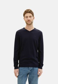 Marineblauer, v-ausschnittiger Strickpullover mit langen Ärmeln. Weicher Stoff, taillierte Passform. Kombiniert mit hellblauen Jeans. Minimale Markenkennzeichnung sichtbar.
