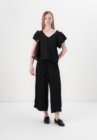Musthave mustriga tekstuuriga top, millel on lühikesed volangvarrukad, koos lõtvade mustade pükste ja mustade mokassiinidega. Mudel kannab burgundia värvi kotti.