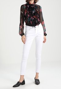 Blusa negra transparente con estampado floral y delantero con volantes, combinada con jeans blancos ajustados y mocasines de cuero negros.