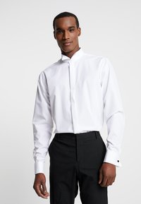 Camicia bianca con colletto alla coreana, maniche lunghe e gemelli neri, abbinata a pantaloni neri. Tessuto morbido e liscio.
