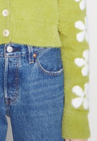 Grön fluffig tröja med vita blommönster på ärmarna, försedda med rundade knappar. Bärs över blå denimjeans med klassisk sömnad.