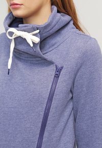 La giacca in pile viola presenta un colletto alto e una cerniera frontale. Ha un laccio bianco e una superficie liscia con dettagli di cucitura visibili.