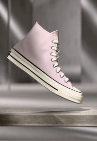 Høye sneakers i lys rosa lerret, med hvite snørebånd, svart stripe på sålen og en teksturert gummitåhette.