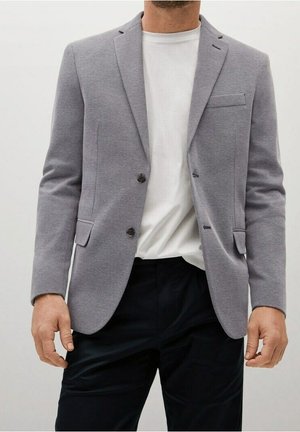 Homme portant un blazer gris clair sur un t-shirt blanc uni et un pantalon noir, debout, les bras détendus le long du corps.