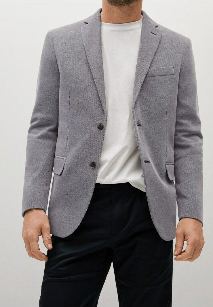Homme portant un blazer gris clair sur un t-shirt blanc uni et un pantalon noir, debout, les bras détendus le long du corps.