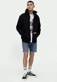 Μαύρο τζάκετ με φερμουάρ από λείο ύφασμα, φορεμένο πάνω από ένα graphic t-shirt. Συνδυασμένο με τζιν σορτς με ρεβέρ, μαύρα αθλητικά παπούτσια και ριγέ κάλτσες.