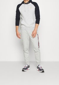 Grauer Kapuzenpullover mit dunkelblauen Ärmeln, Kängurutasche und gelbem Logo. Graue Jogginghose mit "GAP 1969" in kastanienbraun bedruckt. schwarze und graue Laufschuhe.