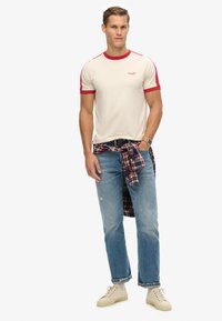 Beige T-shirt med røde accenter, parret med lyseblå jeans. En ternet flannel skjorte bundet om livet og hvide sneakers fuldender looket.