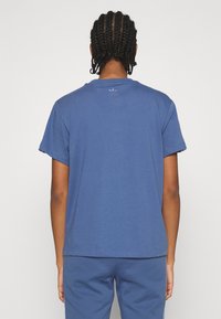 Blå bomullströja med korta ärmar, rund halsringning och en liten logotyp på övre delen av ryggen. Slät textur, vanlig passform och rak nederkant.