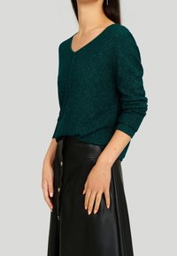 Femme portant un pull vert foncé pailleté à col en V avec manches longues et une jupe en cuir noire boutonnée taille haute, debout de profil.