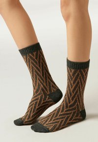 Gestrickte Socken mit einem Zickzackmuster in Orange und Dunkelgrau, ausgestattet mit einem strukturierten gerippten Bund. Das Design hat eine Länge bis zur Mitte der Wade.