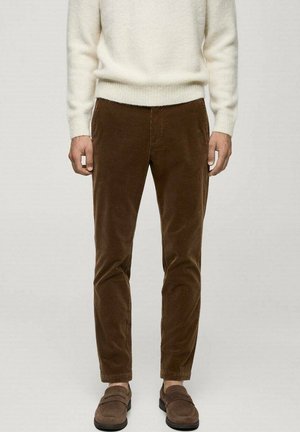 Man draagt bruine corduroy broek, bruine suède loafers en een crèmekleurige gebreide trui, staand tegen een effen achtergrond.
