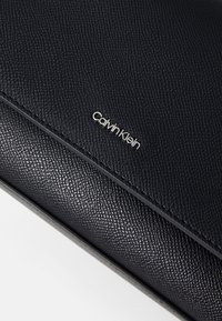 Čierna kožená peňaženka s textúrovaným povrchom, so strieborným logom "Calvin Klein" na prednej strane a prešívaním pozdĺž okraja.