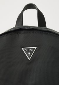 Sac à dos en nylon noir avec une plaque logo triangulaire en argent portant le texte "GUESS USA" et une surface texturée. Poignée supérieure rembourrée.