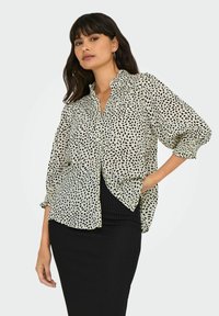 Losse crèmekleurige blouse met zwarte abstracte vlekken, knoopsluiting aan de voorkant en driekwart mouwen, gecombineerd met een zwarte ribstoffen rok.