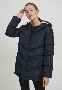 Marineblauer, isolierter Daunenjacke mit Kapuze, ausgestattet mit einem durchgehenden Reißverschluss, seitlichen Taschen und diagonalem Nähmuster. Weiche Innenausstattung sichtbar.