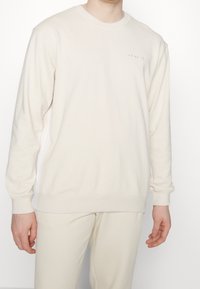 Sudadera de cuello redondo color crema, hecha de un tejido suave, con mangas largas y detalles sutiles de la marca en la parte frontal. Combinada con pantalones a juego.
