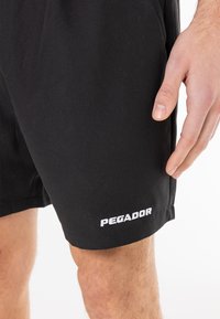Schwarze Sportshorts mit weißem "PEGADOR" Logo am Saum, getragen von einer Person mit locker seitlich herabhängender Hand.