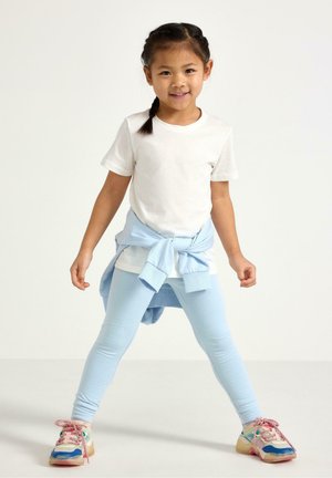 Ung pige står med benene fra hinanden, iført hvid t-shirt, lyseblå leggings, bundet sweatshirt og flerfarvede sneakers, smilende til kameraet.