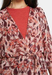 Envueltorio ligero, estampado con diseño de paisley en tonos de rojo y azul marino. Se ata a la cintura, superpuesto a una blusa roja texturizada.