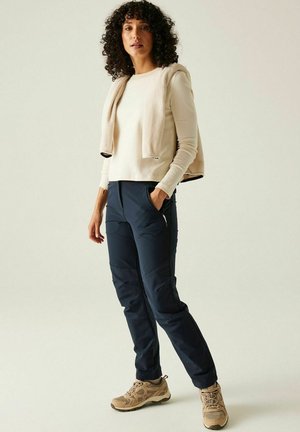 Kobieta o kręconych włosach, ubrana w beżową bluzkę z długim rękawem, beżową kurtkę zarzuconą na ramiona, granatowe spodnie i beżowe buty turystyczne.